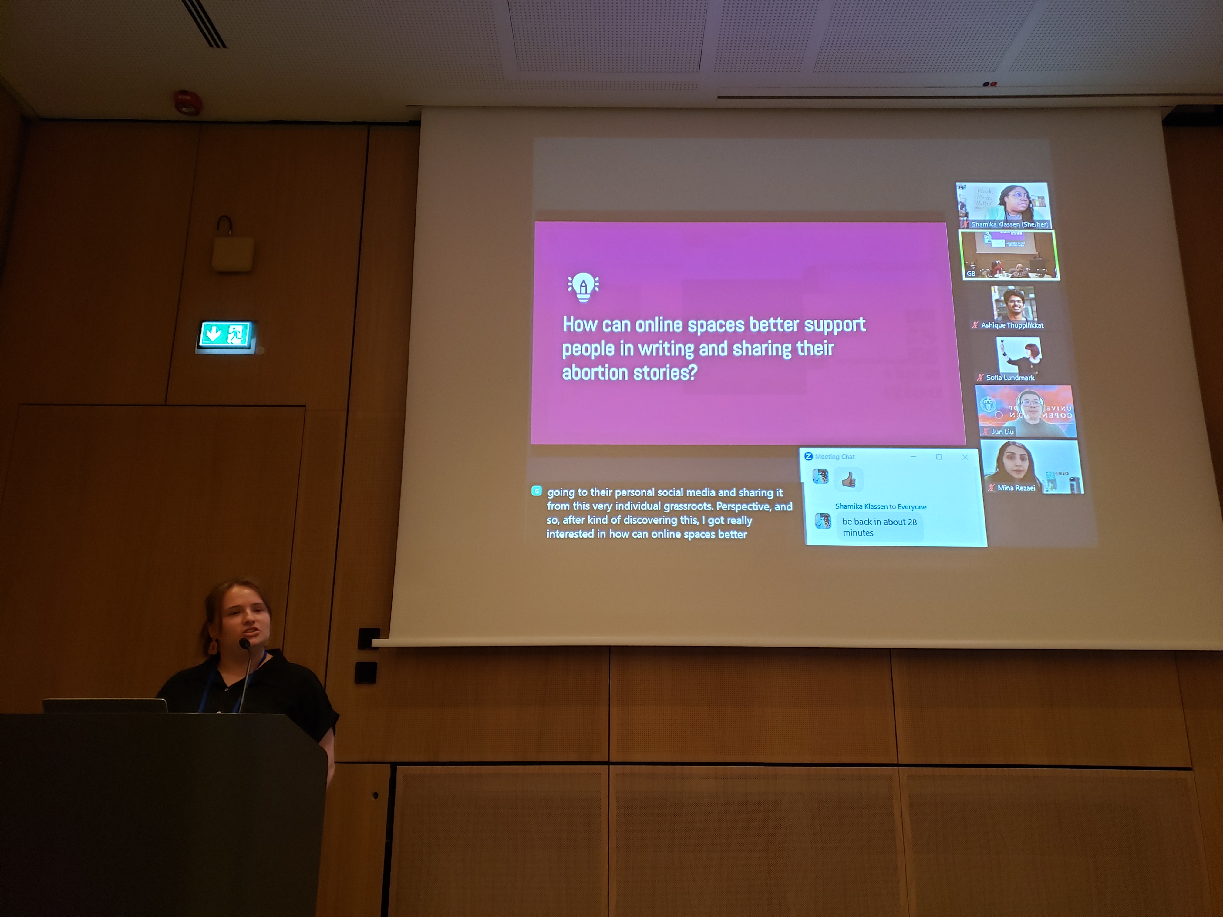 Michaela standing at a podium with a projected screen in the background, giving a hybrid presentation on her abortion storytelling research. The current slide reads "How can online spaces better support people in writing and sharing their abortion stories?"