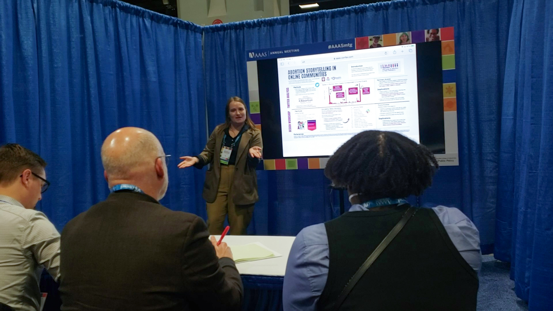 Michaela stands in front of a digital screen displaying her research poster, hands raised in explanation. Three judges sit at a white folding table with their backs to the camera, watching Michaela present. You can't tell in the photo, but Michaela is shouting because the expo hall was very loud.